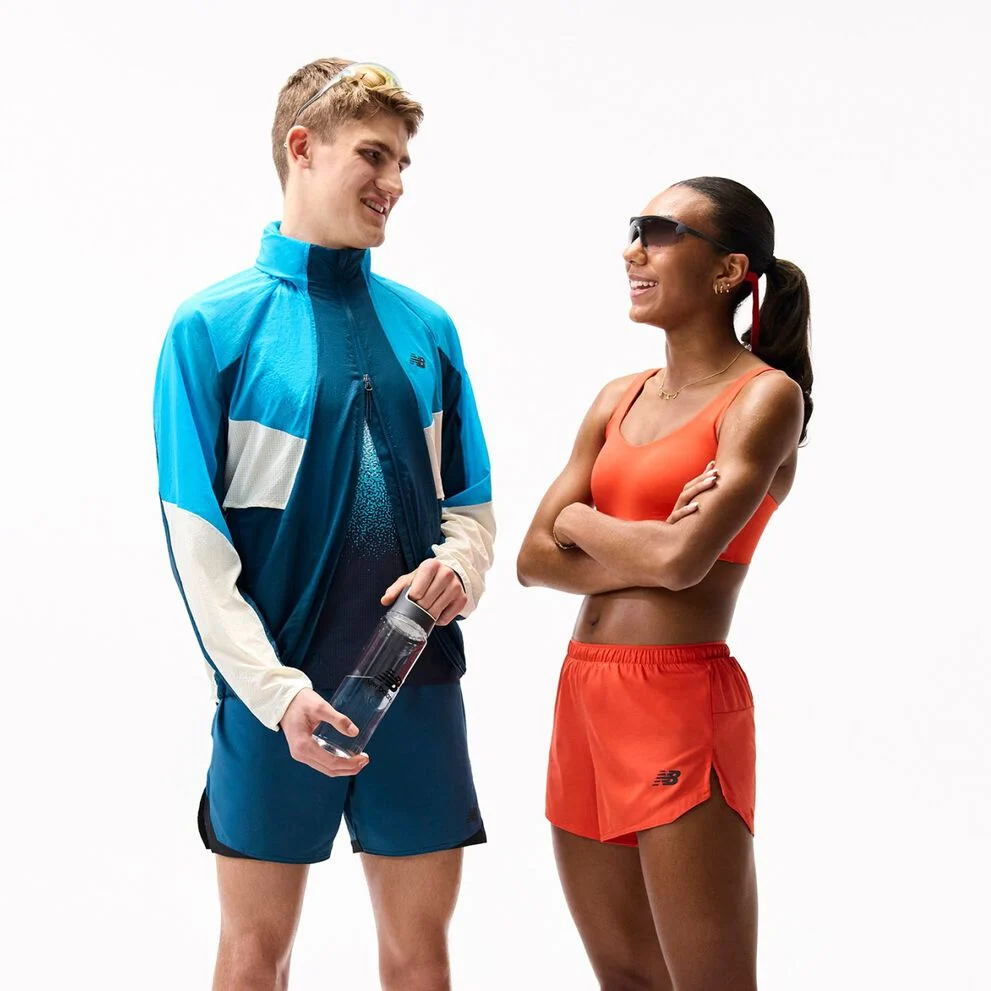 Person in bright orange running outfit and sneakers stands on a white background holding a towel and a water bottle, with sunglasses hanging around the neck.
